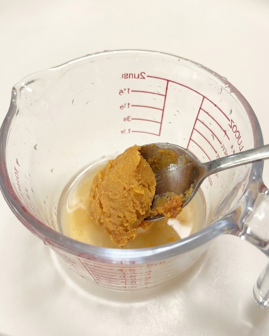 Una cucharada de pasta de miso siendo añadida a una jarra medidora con condimentos líquidos.