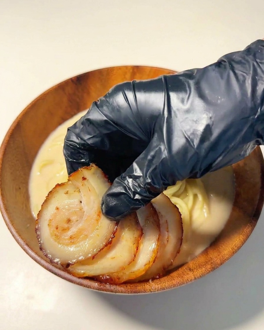 Una mano con guante negro colocando rodajas de cerdo chashu sellado sobre los fideos.