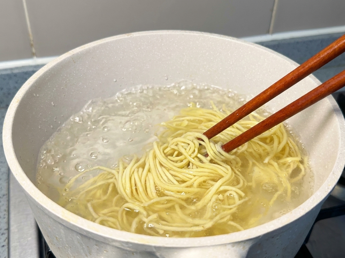 Fideos ramen amarillos frescos siendo levantados con palillos de una olla con agua hirviendo.