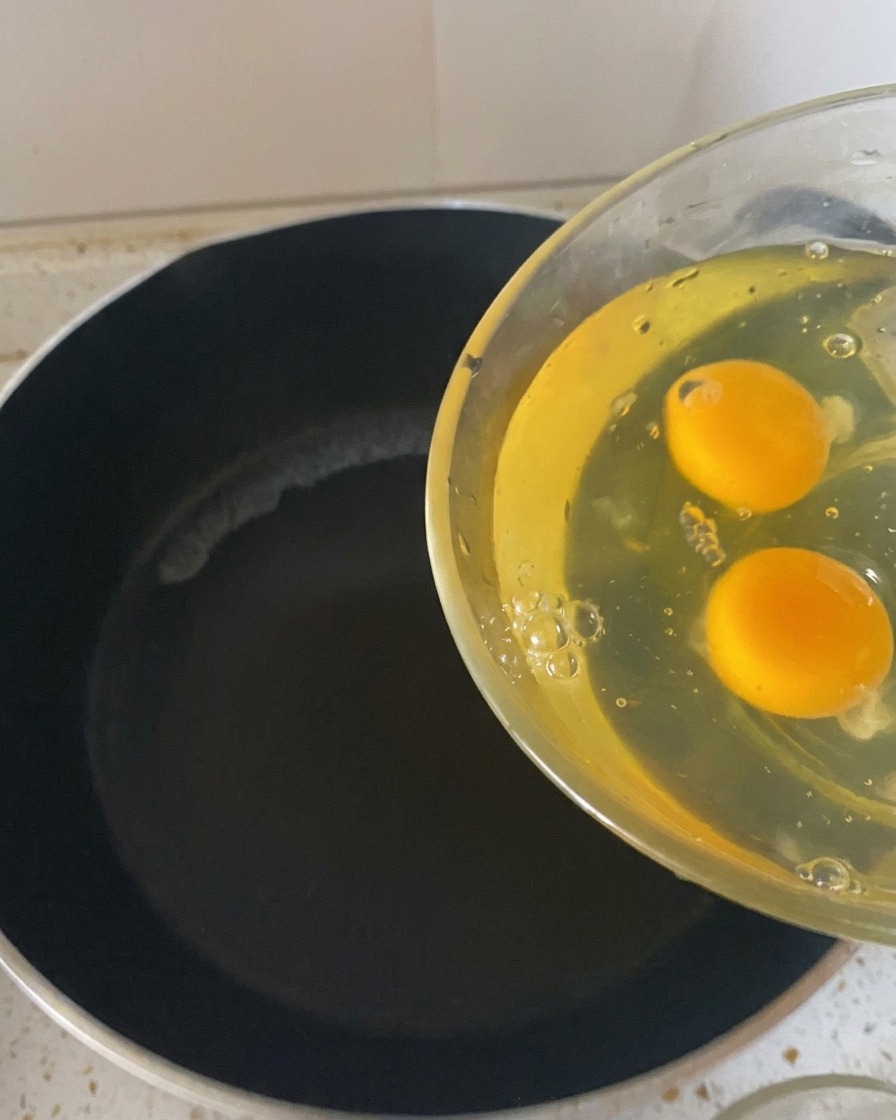 Dos huevos crudos vertiéndose desde un cuenco de cristal transparente a una sartén oscura y caliente.
