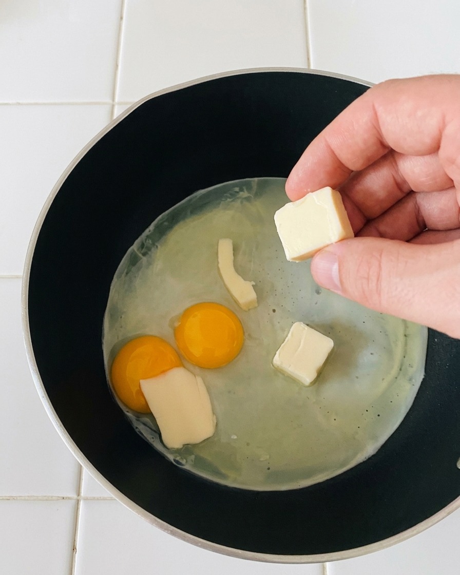 Una mano colocando pequeñas rebanadas rectangulares de queso mozzarella blanco sobre unos huevos fritos cocinándose a fuego lento.