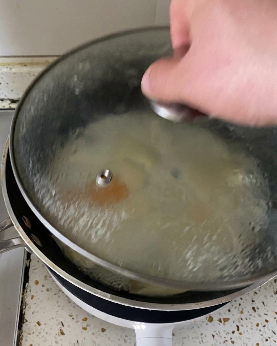 Una mano levantando la tapa de cristal de una sartén, dejando ver el vapor y unos huevos perfectamente cocinados cubiertos de queso fundido.