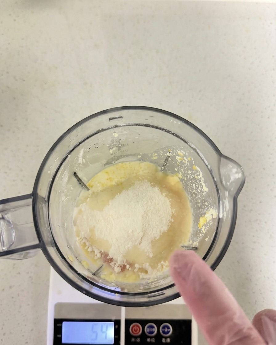 Harina y otros ingredientes secos sobre la mezcla húmeda dentro del vaso de la licuadora sobre una báscula de cocina.
