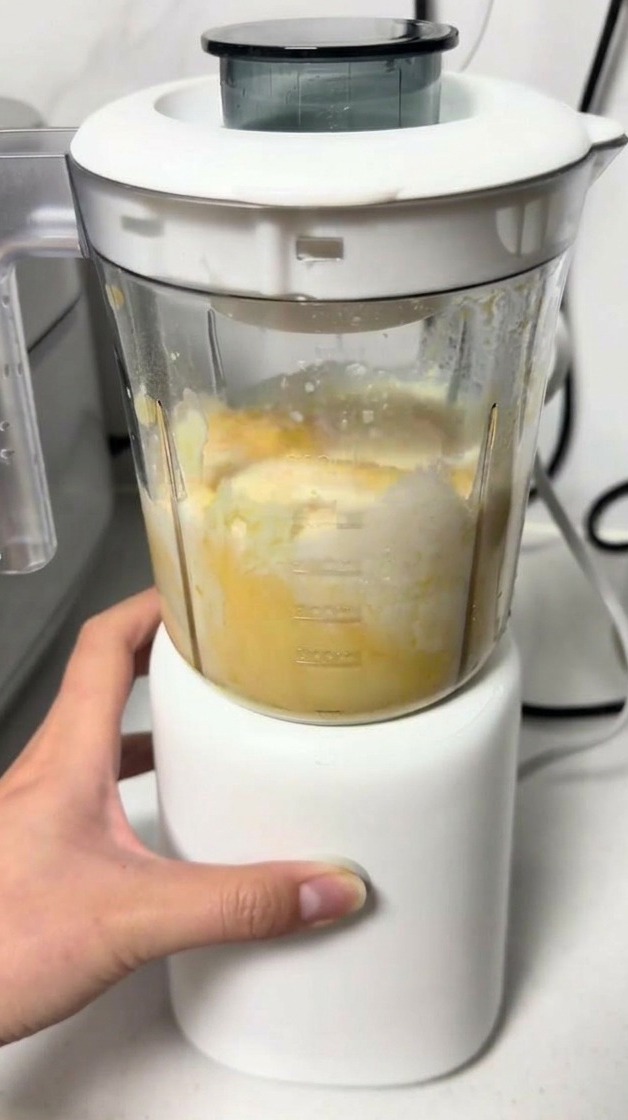 Una mano operando una pequeña licuadora mientras mezcla una masa amarilla de queso y huevo.