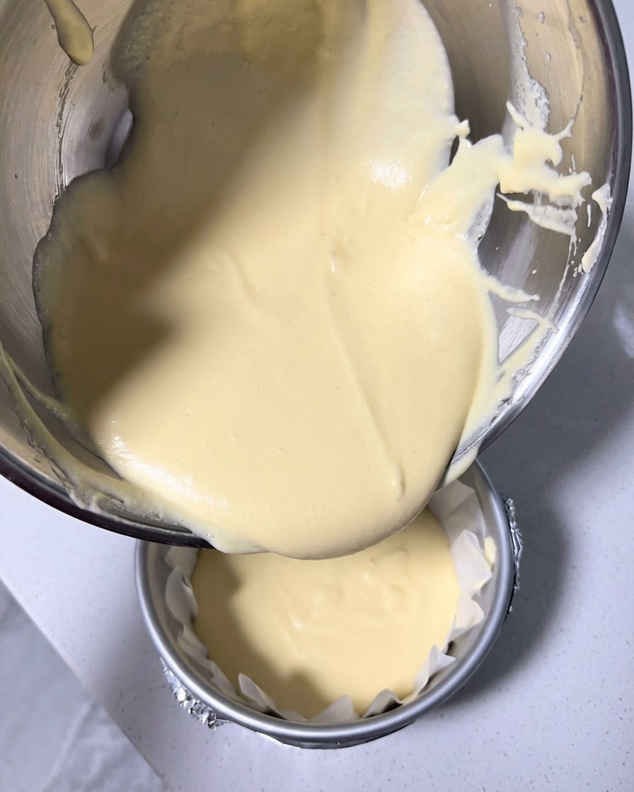 Masa de pastel de queso amarilla pálida siendo vertida en un molde redondo forrado con papel de hornear arrugado.