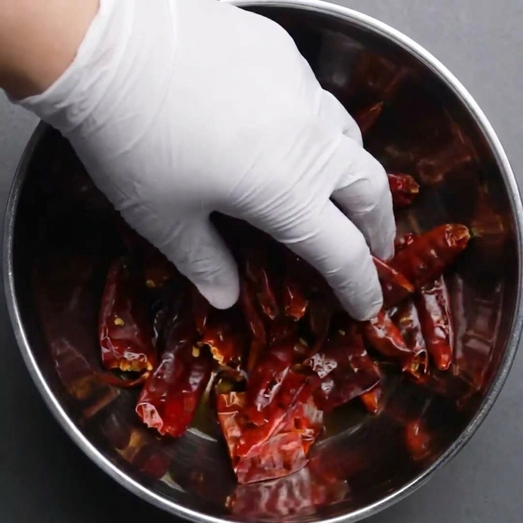 Una mano con guante sumergiendo chiles rojos secos en un bol con agua.