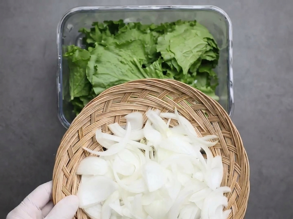 Tiras de cebolla blanca cayendo sobre una cama de lechuga verde troceada en un recipiente de vidrio.