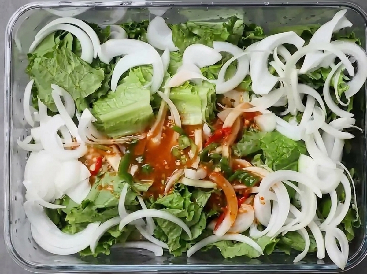 Caldo de chile rojo vertido sobre lechuga fresca y cebolla en un recipiente rectangular de vidrio.