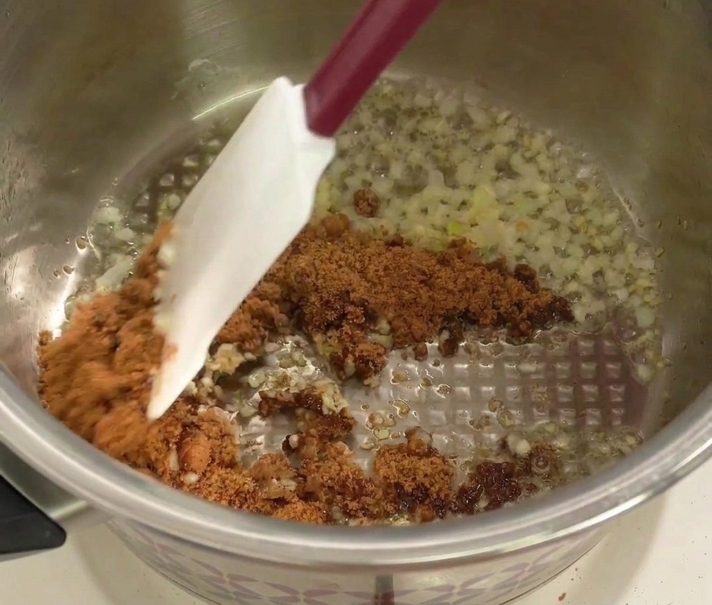 Una espátula mezclando azúcar moreno con el sofrito de cebolla y ajo.
