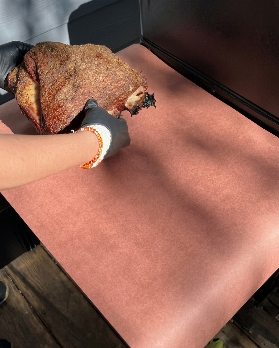 Manos con guantes sosteniendo un costillar ahumado con costra oscura, colocándolo sobre papel de carnicero rosa.