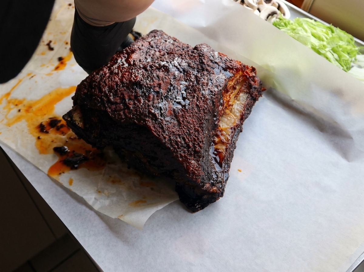 Manos con guantes negros colocando una costilla de res asada y oscura sobre una hoja de papel para hornear blanco.