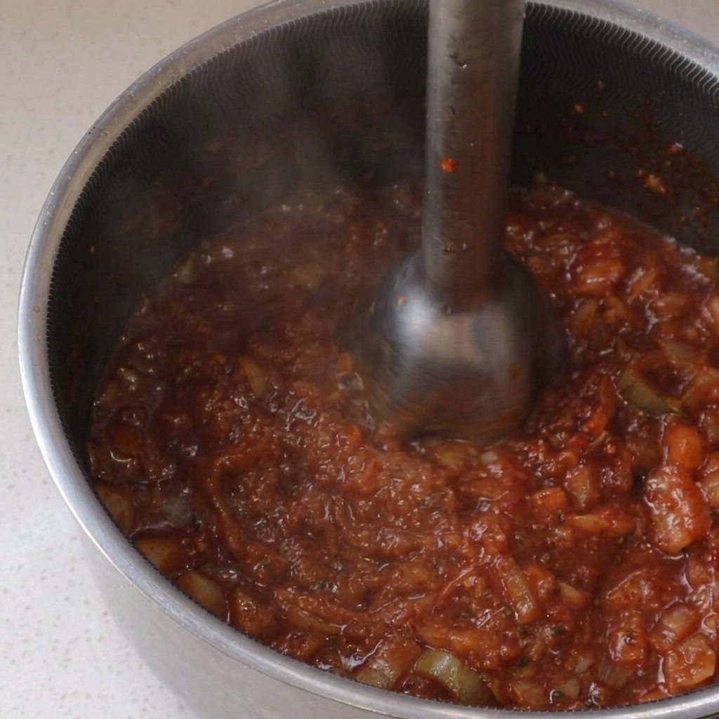 Uso de una batidora de mano para triturar una salsa BBQ roja espesa dentro de una olla de acero inoxidable.