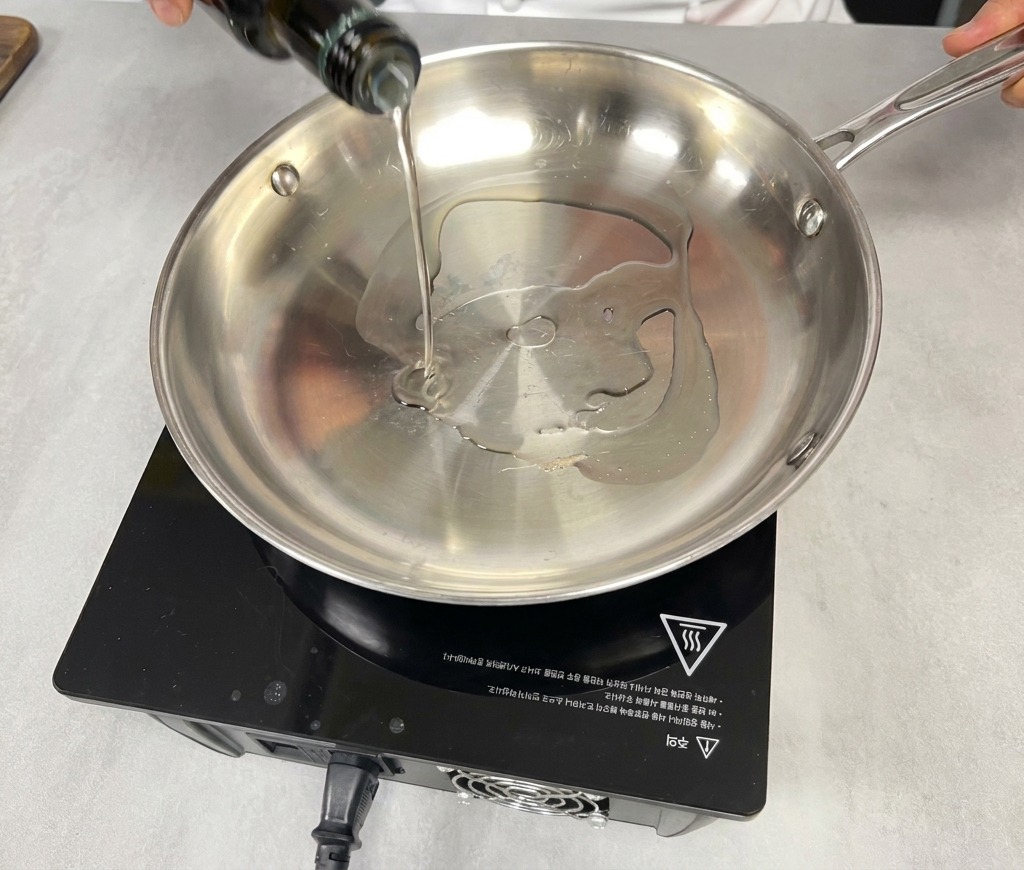 Una mano vertiendo aceite de cocina de una botella de vidrio verde oscuro en una sartén vacía de acero inoxidable sobre una estufa de inducción negra.