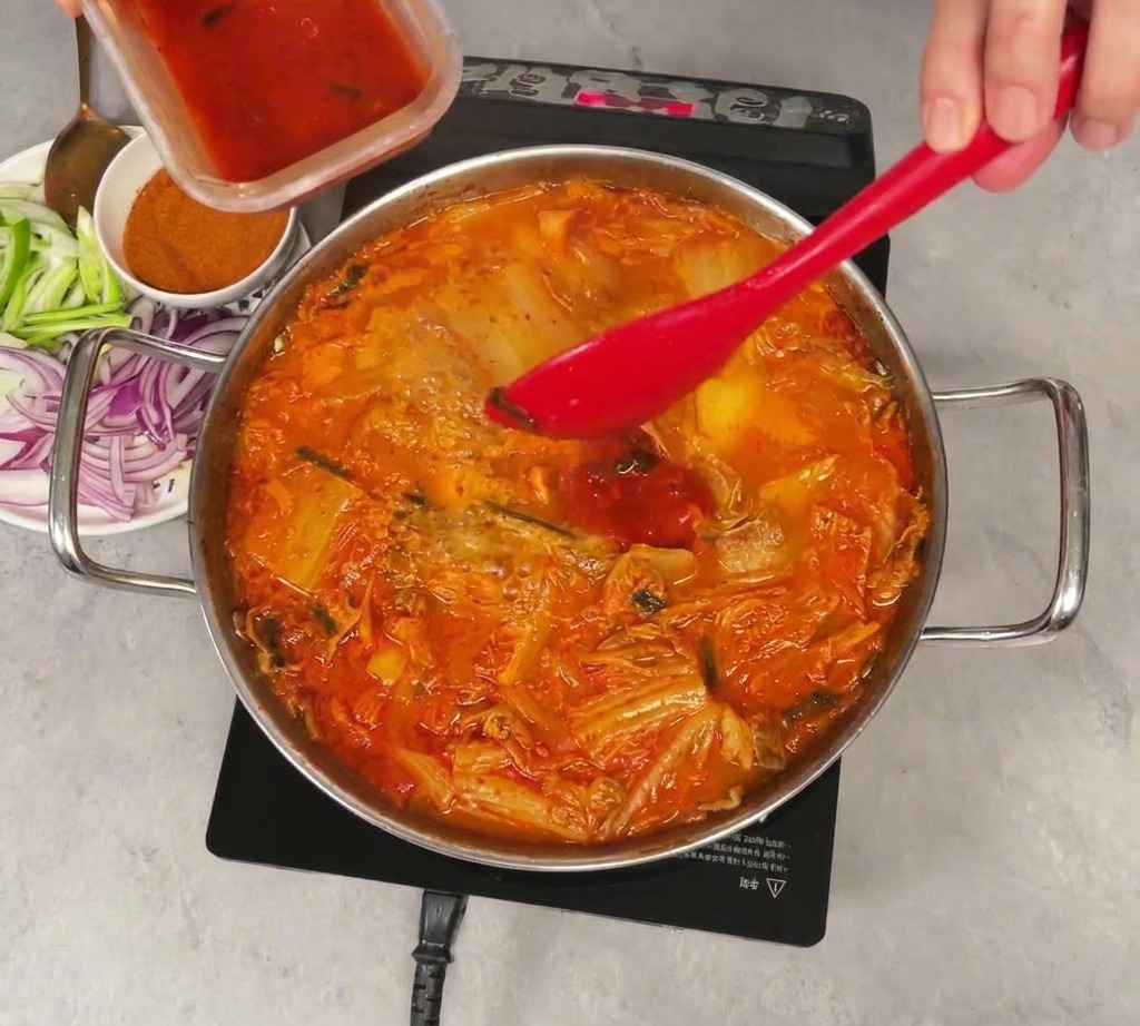 Jugo rojo brillante de kimchi siendo vertido de un recipiente de plástico pequeño en una olla a fuego lento con estofado de kimchi, mezclado con una espátula roja.