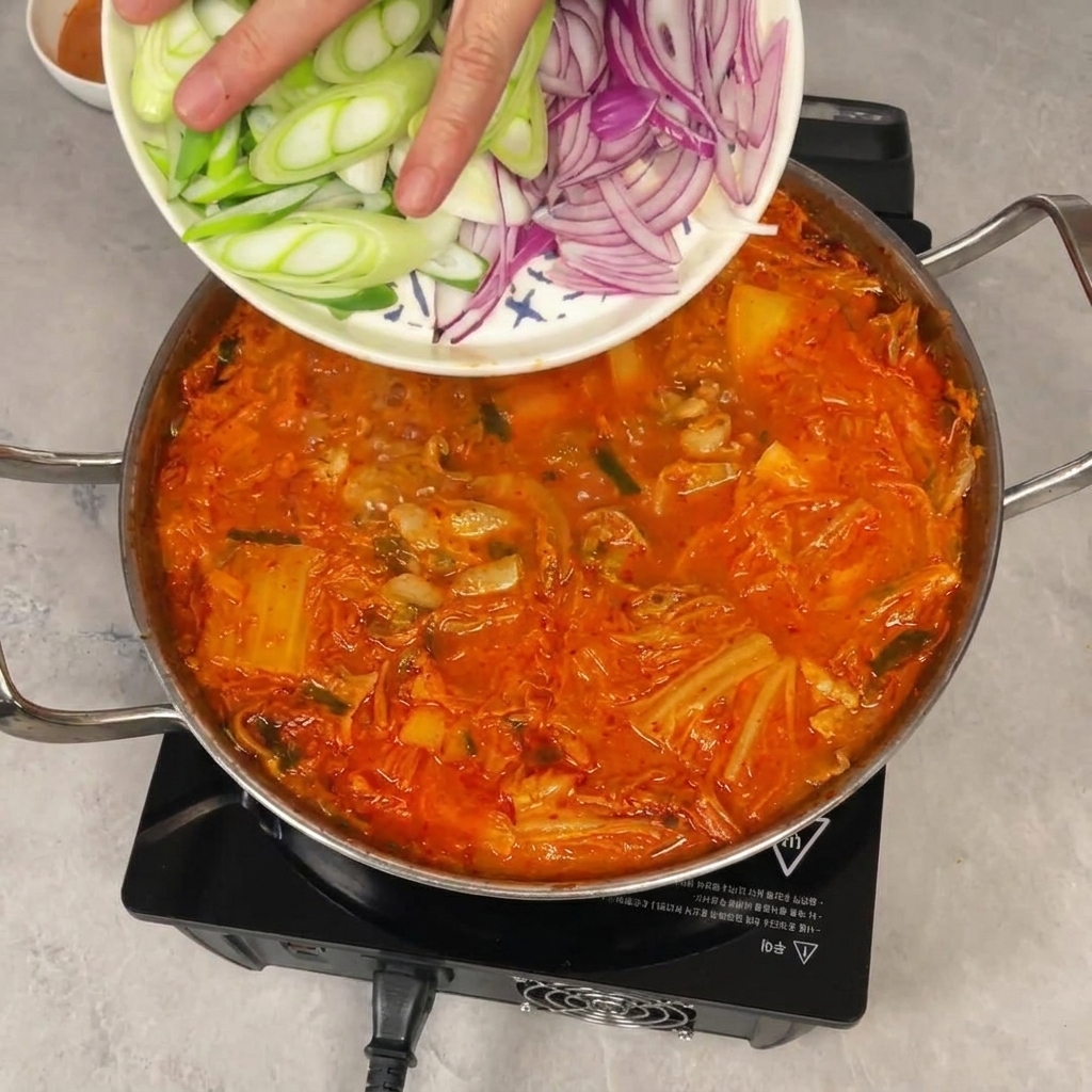 Una mano deslizando cebolleta y cebolla morada picadas desde un plato hacia una olla burbujeante de estofado de kimchi.