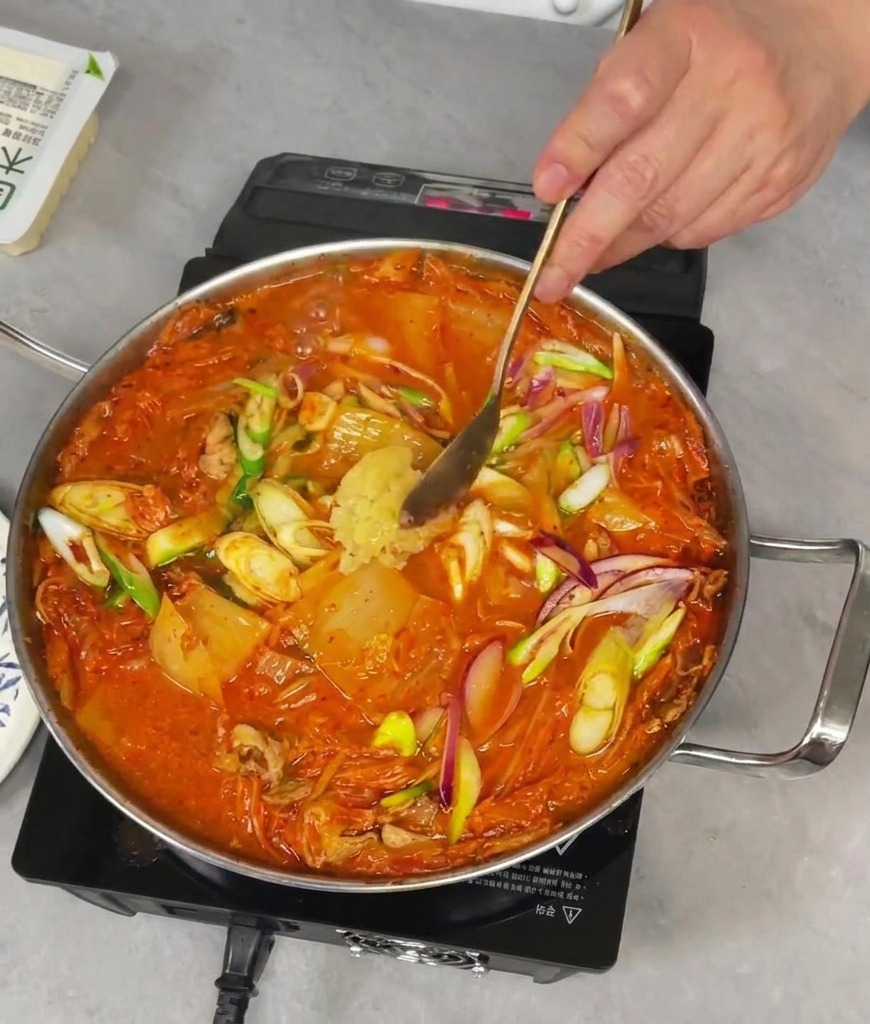 Añadiendo una cucharada de ajo fresco picado a una sartén burbujeante de estofado rojo de kimchi.