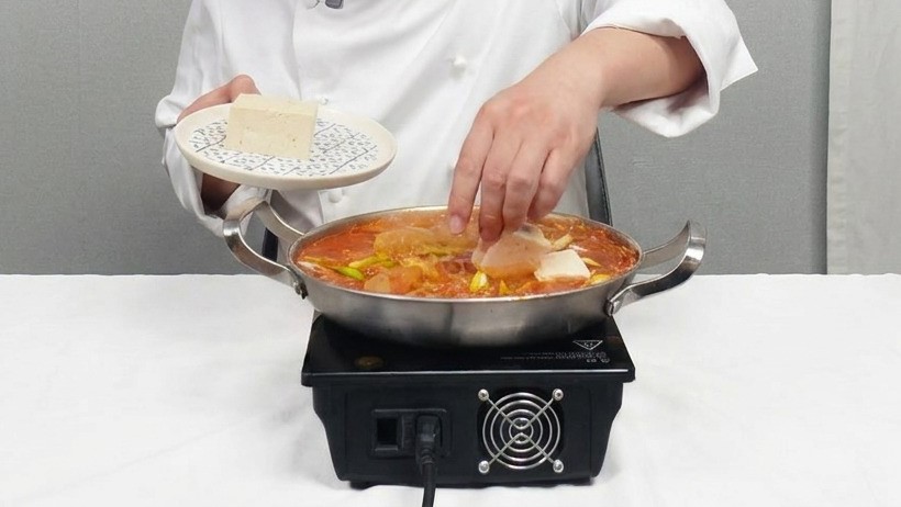 Un chef colocando suavemente rodajas cuadradas de tofu blanco en una olla a fuego lento de estofado de kimchi.
