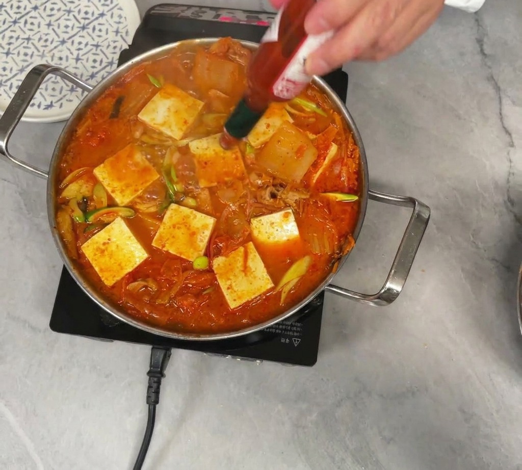 Una persona vertiendo unas gotas de salsa picante Tabasco en una olla hirviendo de estofado de kimchi cubierto con piezas cuadradas de tofu blanco.