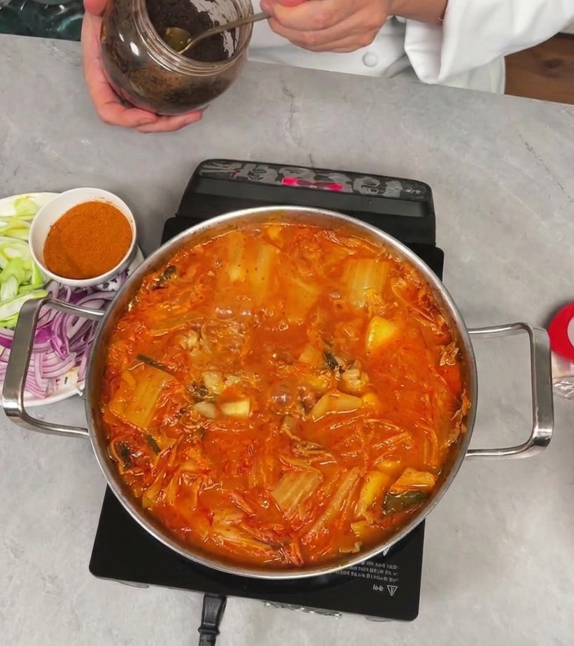 Un chef sosteniendo un frasco y sacando una cucharada de pasta de soja oscura y espesa para añadir al estofado de kimchi.