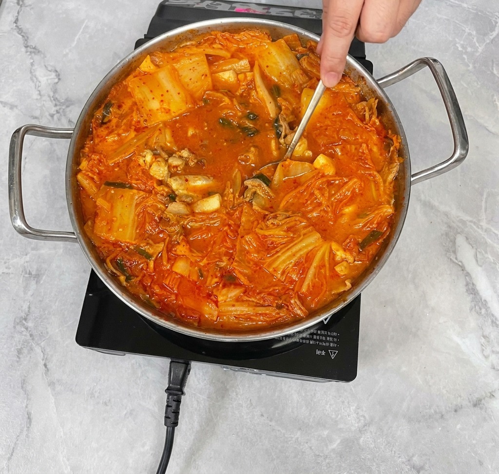 Una mano usando una cuchara de acero inoxidable para remover suavemente un vibrante estofado de kimchi, mezclando los condimentos en el caldo.