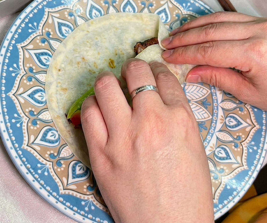 Dos manos enrollando cuidadosamente una tortilla rellena de ternera y vegetales.
