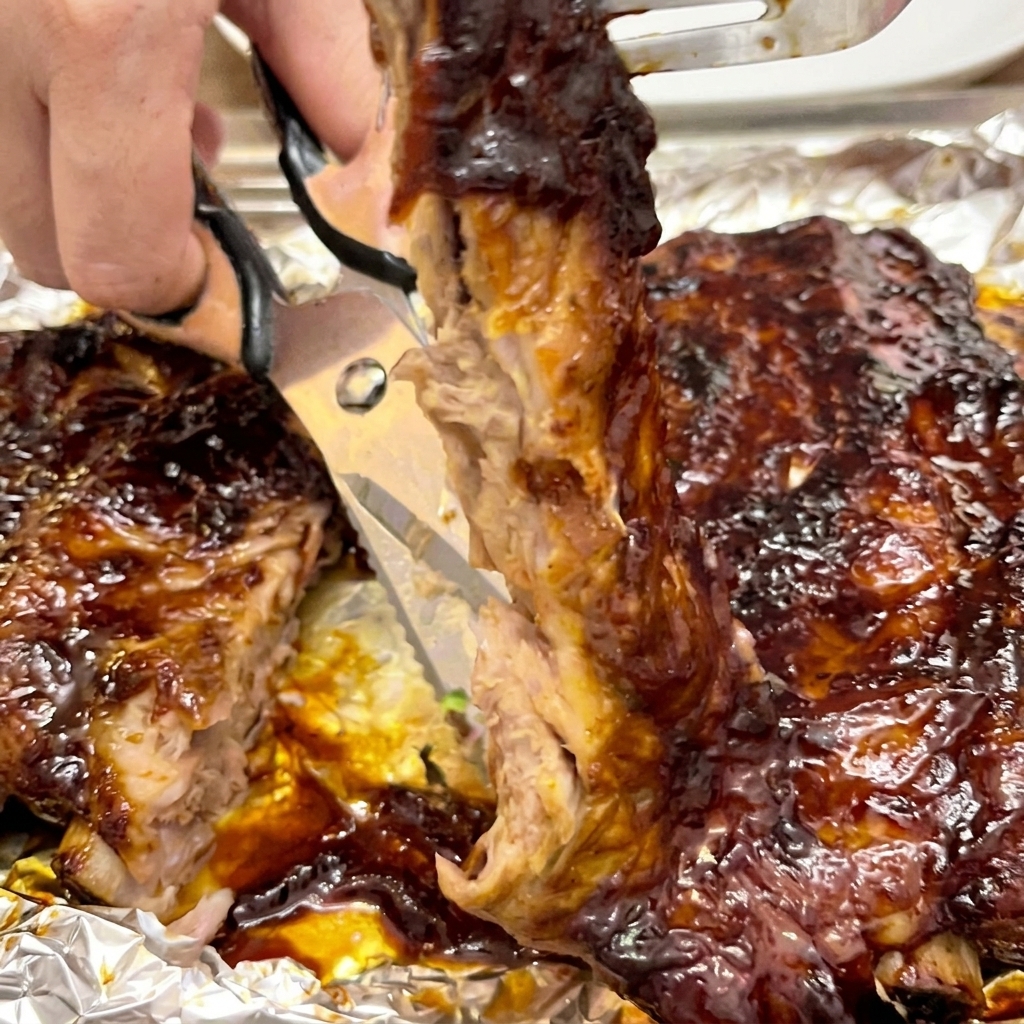 Primer plano de unas tijeras de cocina cortando entre los huesos de unas costillas tiernas cubiertas de salsa.