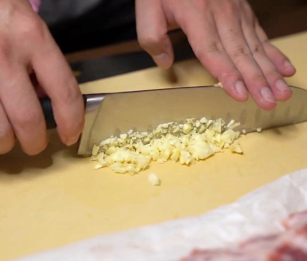 Manos picando finamente dientes de ajo frescos con un cuchillo de chef sobre una tabla de cortar amarilla.