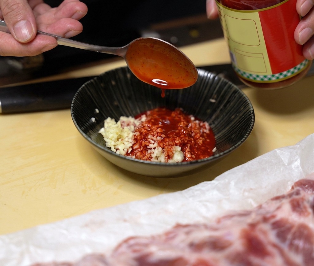 Una cucharada de líquido rojo brillante de tofu fermentado siendo añadida a un bol negro con ajo picado.