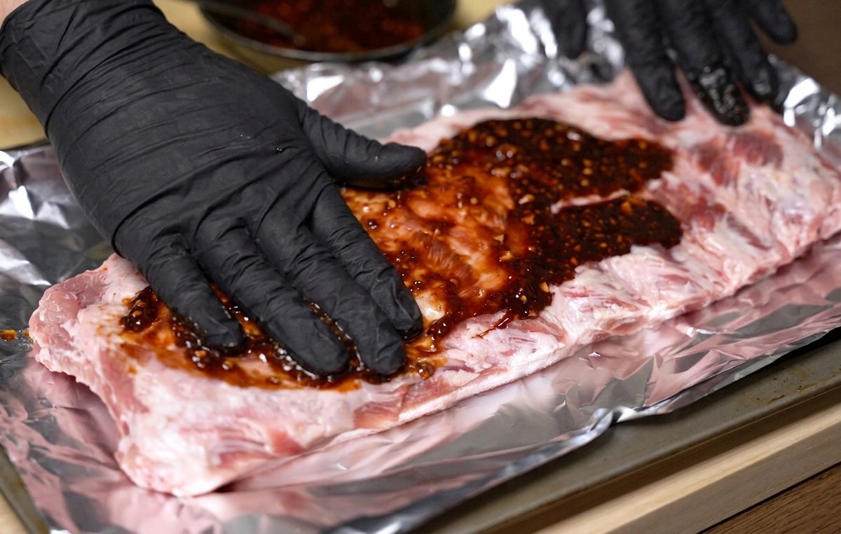 Manos con guantes frotando un marinado espeso de color rojo oscuro sobre un costillar de cerdo sobre papel de aluminio.