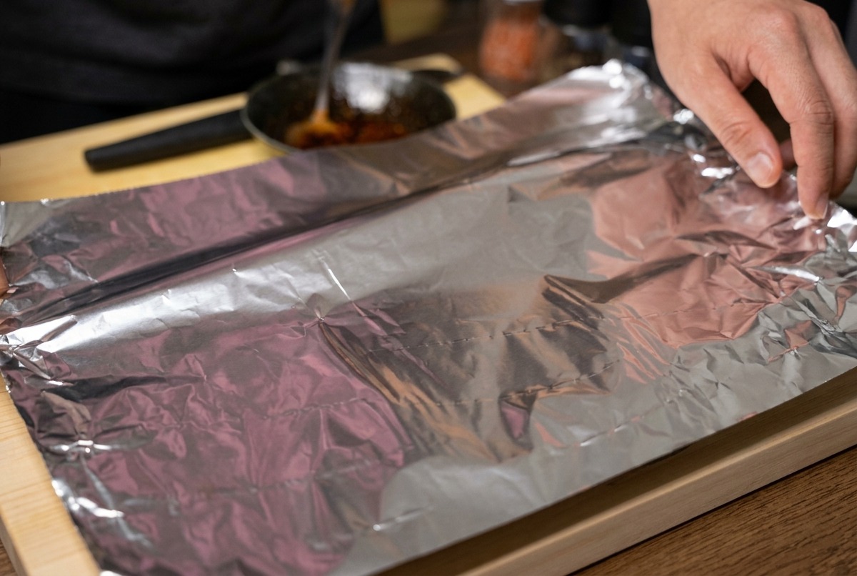 Manos doblando una hoja grande de papel de aluminio para envolver herméticamente un costillar de cerdo marinado.