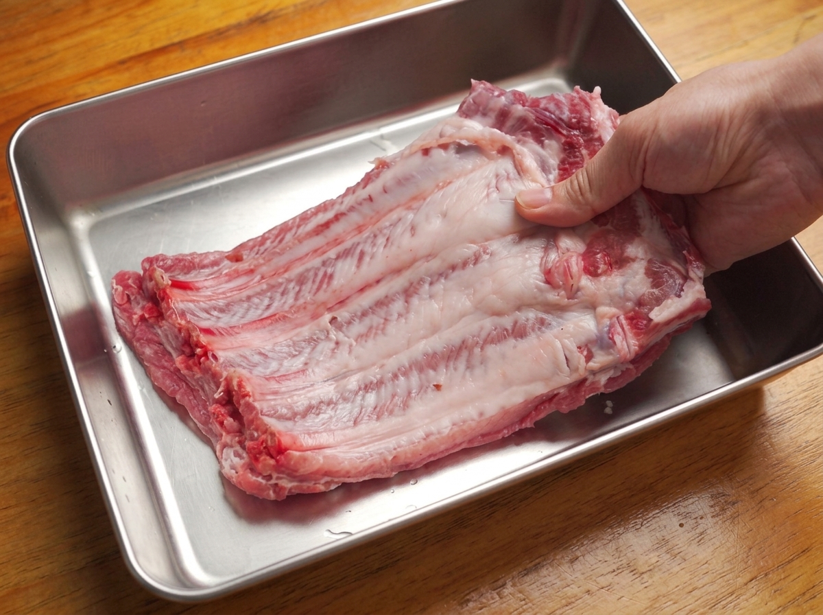 Manos retirando la fina membrana de la parte posterior de un costillar de cerdo crudo en una bandeja metálica.
