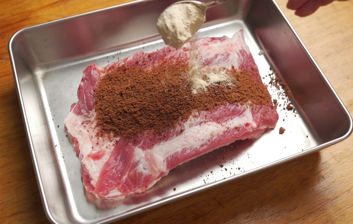 Una bandeja con costillas de cerdo crudas siendo espolvoreadas con azúcar morena y una mezcla de especias secas desde una cuchara.