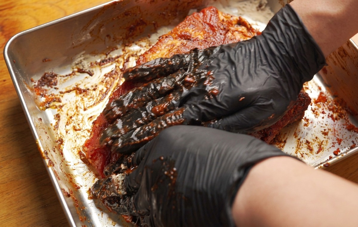 Manos con guantes frotando firmemente una pasta de marinado marrón rojizo en un costillar de cerdo en una bandeja de metal.