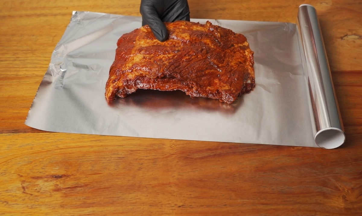 Un costillar de cerdo marinado siendo colocado sobre una hoja limpia de papel de aluminio en una mesa de madera.