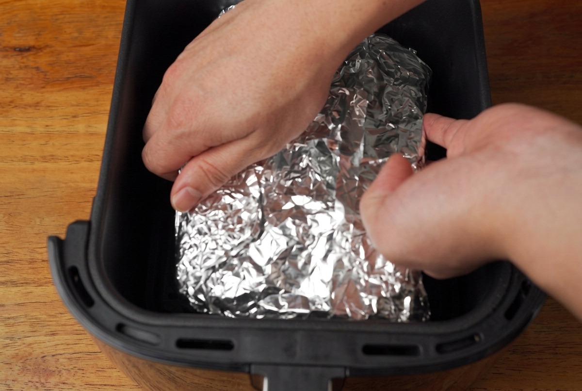 Manos colocando un paquete envuelto en papel de aluminio que contiene costillas de cerdo en la canasta negra de una freidora de aire.