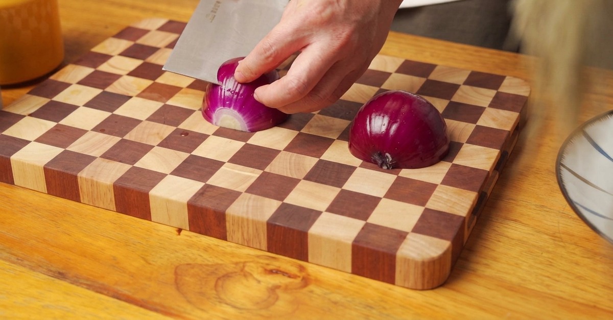 Manos usando un hacha de cocina para picar media cebolla roja en una tabla de cortar de madera.