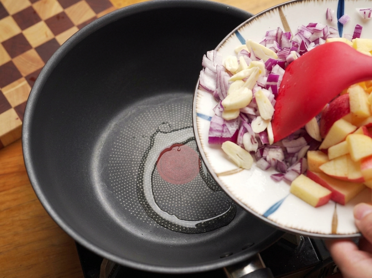 Usando una espátula para deslizar cebolla roja picada y ajo desde un plato hacia una sartén caliente con aceite.