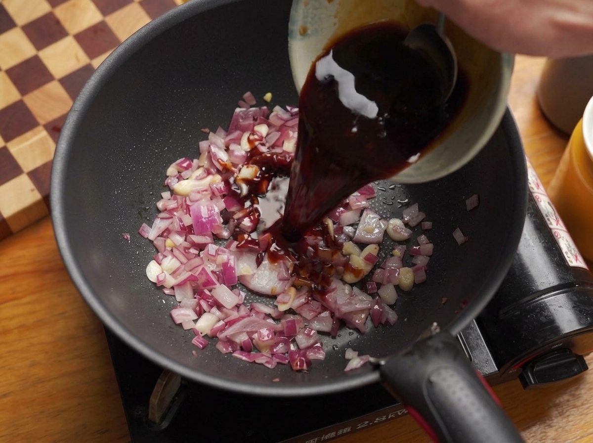 Vertiendo una mezcla de salsa líquida oscura y salada desde un bol hacia una sartén con cebollas y ajo sofritos.