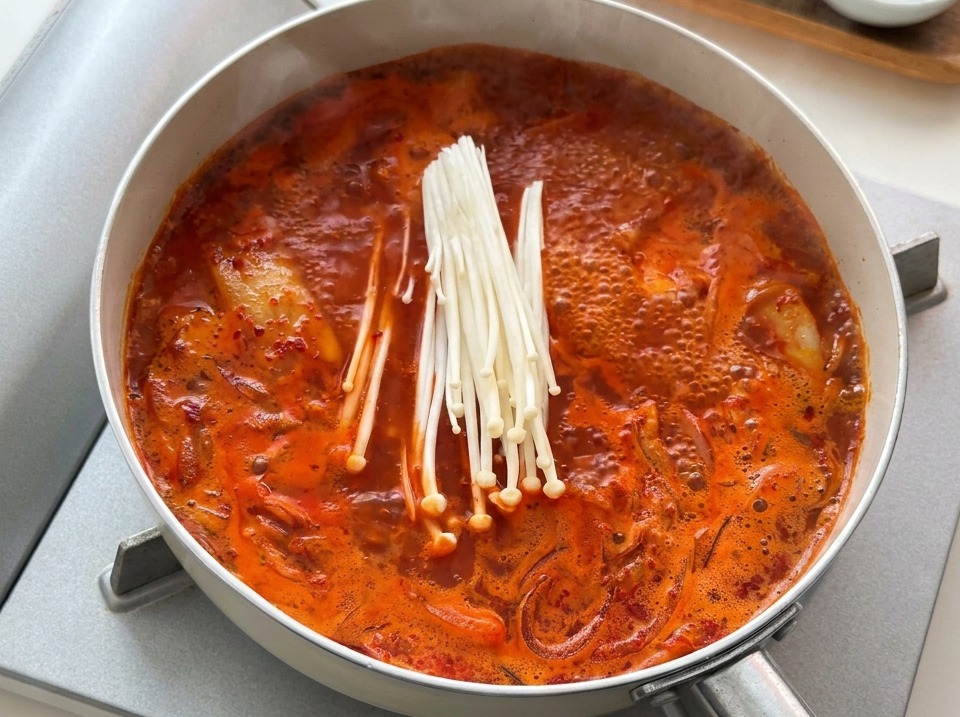 Un manojo de setas enoki blancas crudas añadiéndose a una sartén de estofado de kimchi rojo hirviendo.