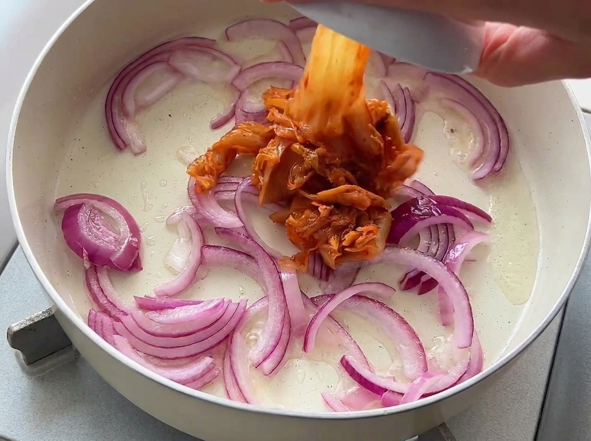 Kimchi picado añadiéndose desde un pequeño bol a una sartén con cebollas rojas sofritas.