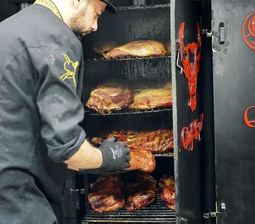 Chef retirando una pieza grande de res ahumada con una costra oscura de un ahumador industrial