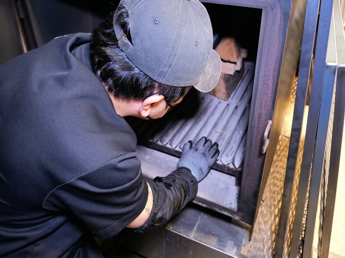 Una persona colocando troncos de madera dentro de la caja de fuego de un gran ahumador de metal.