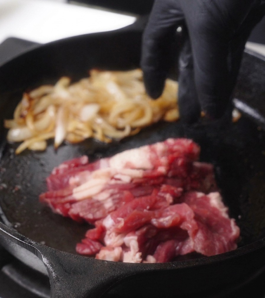 Mano añadiendo láminas de carne roja a la sartén caliente junto a las cebollas ya cocinadas.