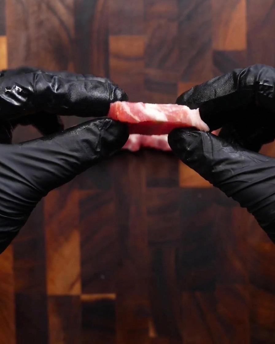 Dos manos con guantes negros sosteniendo una sola rebanada de panceta de cerdo cruda recién cortada para mostrar su grosor, similar al de una moneda.