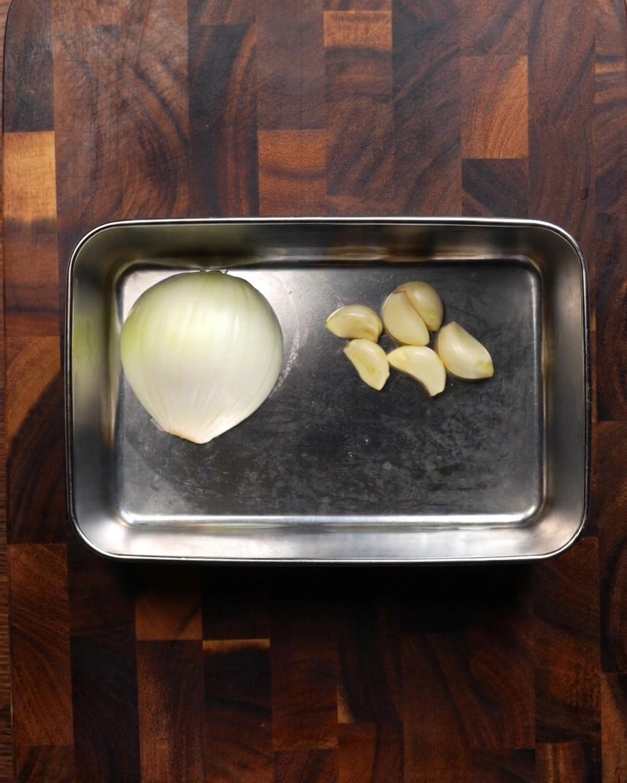 Una bandeja de acero inoxidable que sostiene la mitad de una cebolla blanca y varios dientes de ajo pelados sobre una tabla de madera.