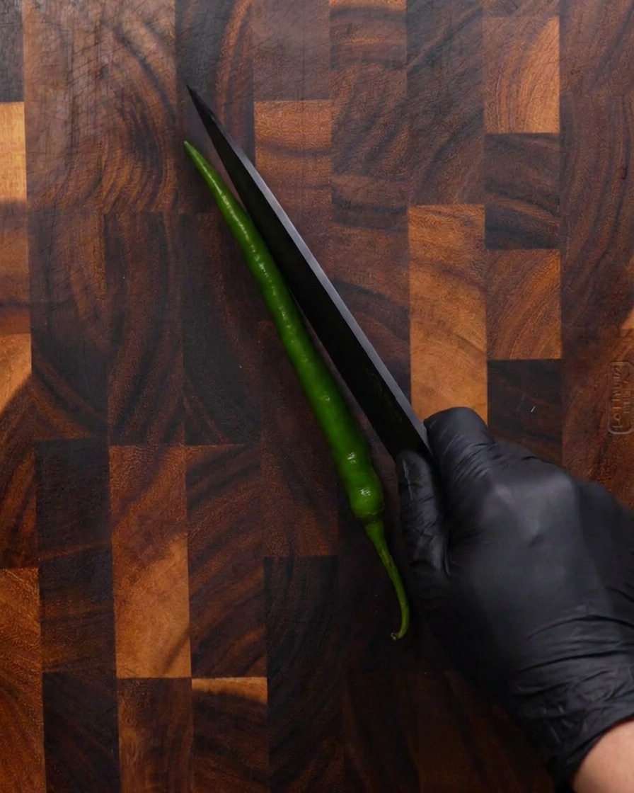 Una mano con un guante negro usando un cuchillo afilado negro para cortar un chile verde largo en diagonal sobre una tabla de cortar de madera.