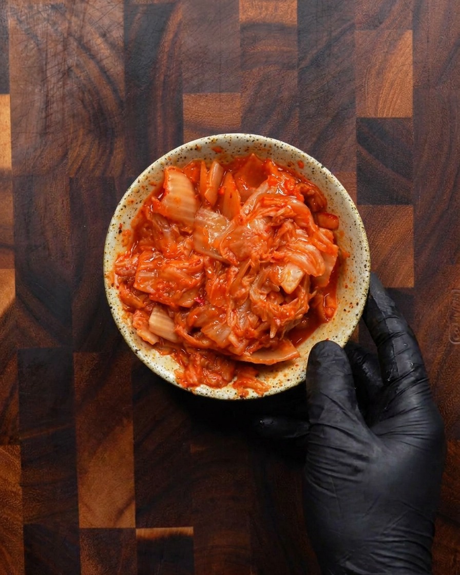Una mano enguantada que sostiene un pequeño tazón rústico moteado lleno hasta el borde con kimchi fermentado picado de color rojo brillante.
