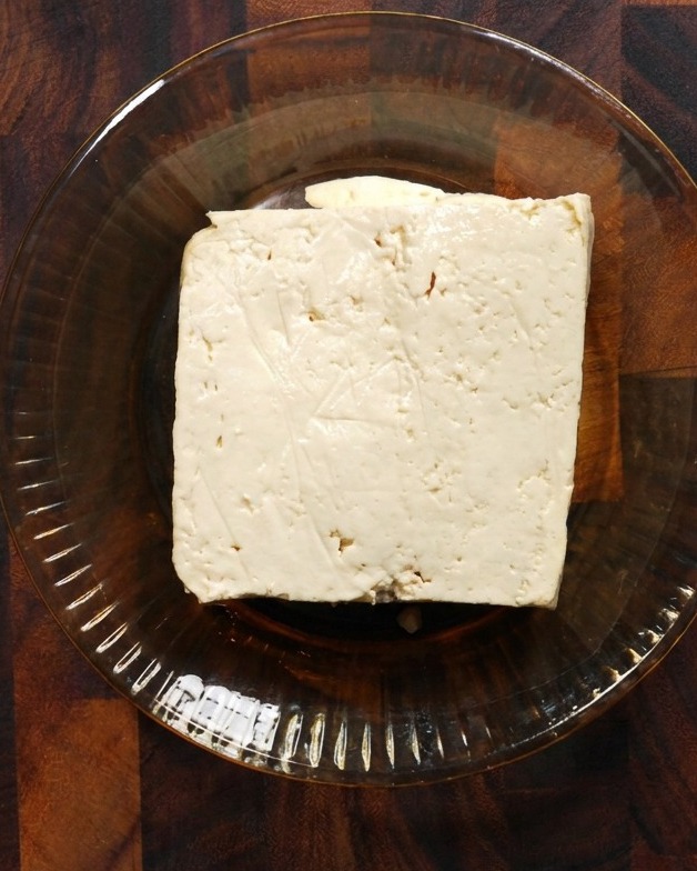 Un bloque cuadrado de tofu blanco firme descansando en un recipiente de vidrio poco profundo sobre una superficie de madera.