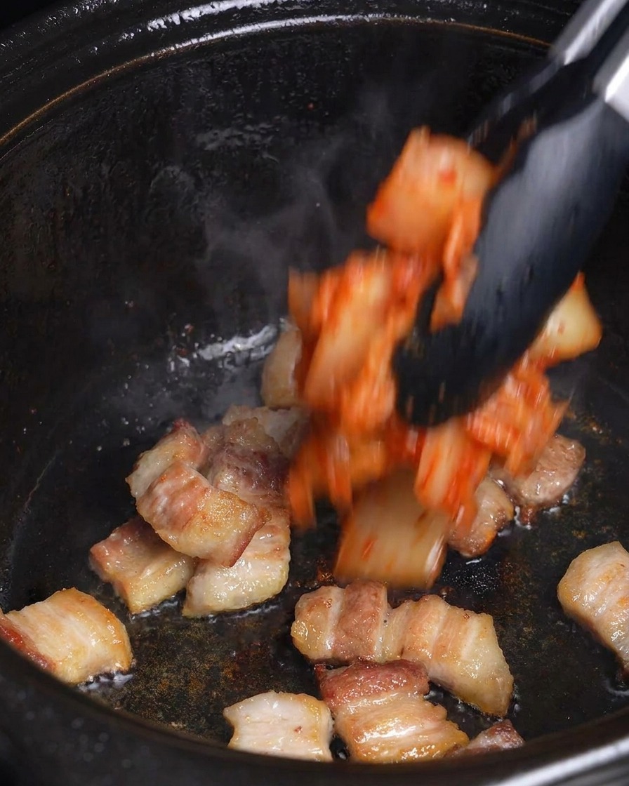 Kimchi rojo picado que se agrega a la panceta de cerdo dorada en una olla negra con unas pinzas.