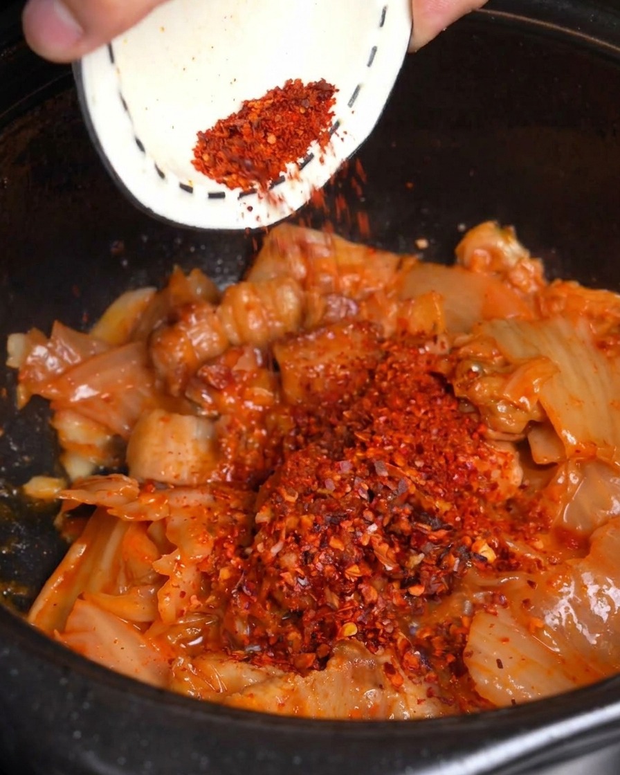 Chile rojo en polvo vertiéndose desde un tazón pequeño sobre el kimchi y la panceta salteados en una olla negra.